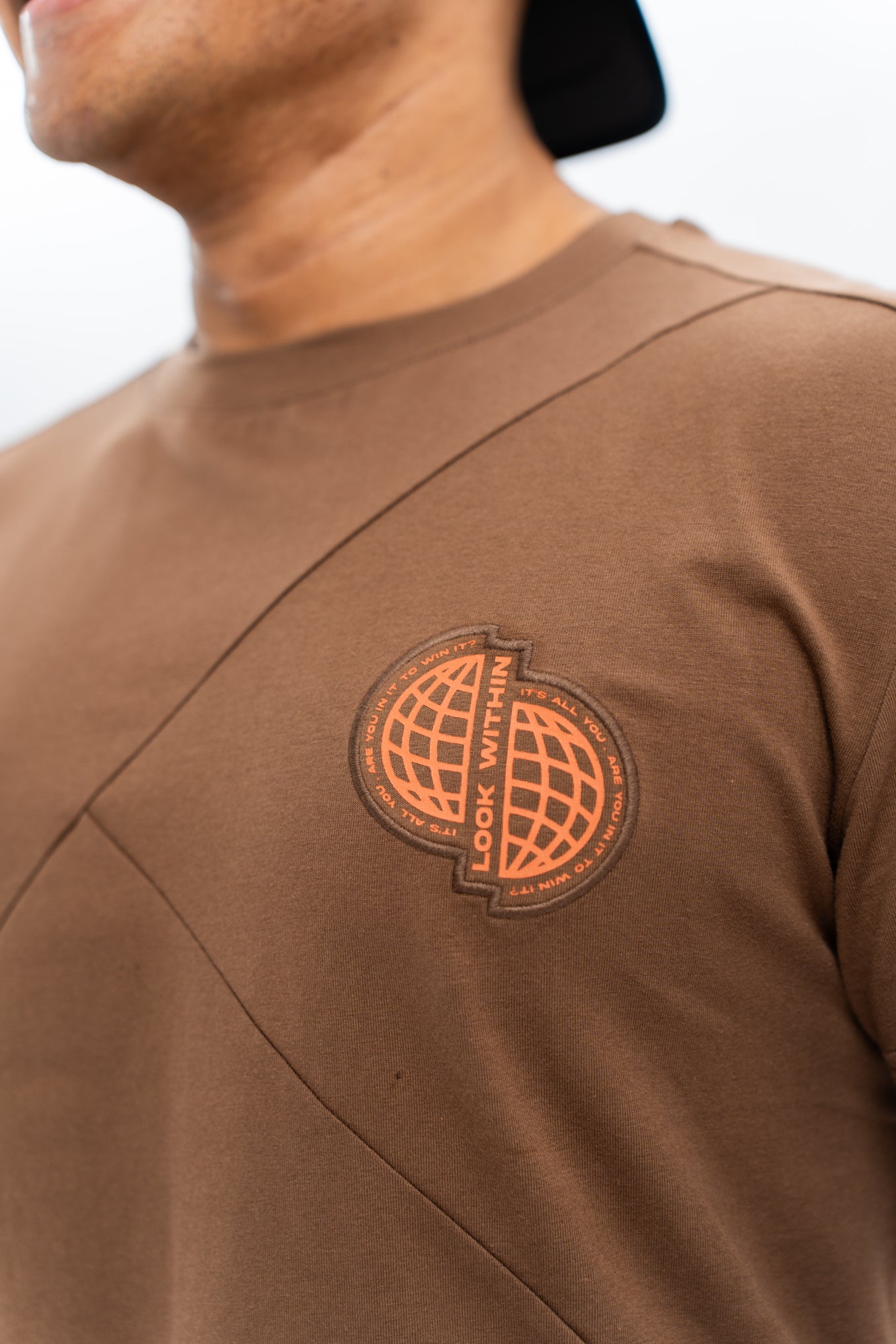 Close-up of a brown oversized tee with a circular brand logo in orange and white on the chest. Cozterra Zoda.sg